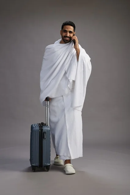 A portrait of an Arab Saudi Gulf man in Ihram clothing with a travel bag, using a mobile phone, going to Mecca for Hajj and Umrah, full body, gray background.