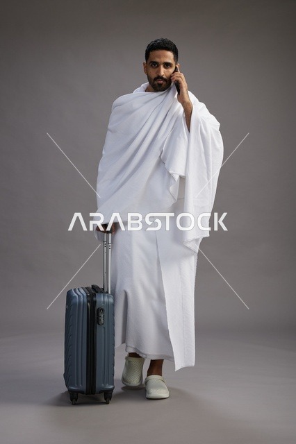 بورتريه لرجل عربي  سعودي خليجي بملابس الإحرام مع حقيبة سفر, استخدام الهاتف المحمول, الذهاب الى مكه للحج والعمره, كامل الجسم, خلفيه رماديه