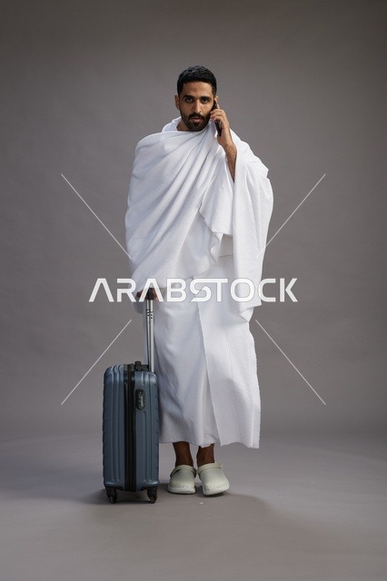 بورتريه لرجل عربي  سعودي خليجي بملابس الإحرام مع حقيبة سفر, استخدام الهاتف المحمول, الذهاب الى مكه للحج والعمره, كامل الجسم, خلفيه رماديه
