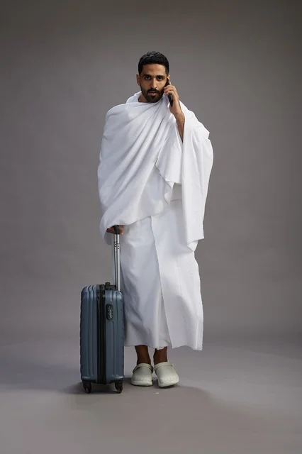 A portrait of an Arab Saudi Gulf man in Ihram clothing with a travel bag, using a mobile phone, going to Mecca for Hajj and Umrah, full body, gray background.