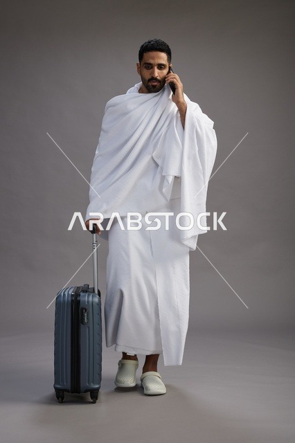 بورتريه لرجل عربي  سعودي خليجي بملابس الإحرام مع حقيبة سفر, استخدام الهاتف المحمول, الذهاب الى مكه للحج والعمره, كامل الجسم, خلفيه رماديه