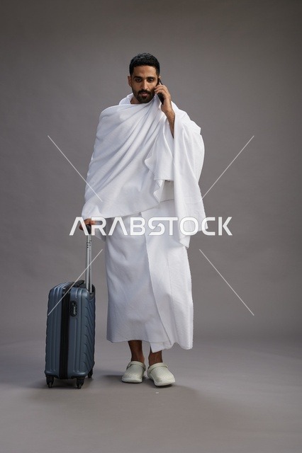 بورتريه لرجل عربي  سعودي خليجي بملابس الإحرام مع حقيبة سفر, استخدام الهاتف المحمول, الذهاب الى مكه للحج والعمره, كامل الجسم, خلفيه رماديه