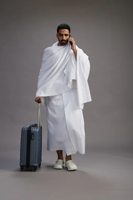 A portrait of an Arab Saudi Gulf man in Ihram clothing with a travel bag, using a mobile phone, going to Mecca for Hajj and Umrah, full body, gray background.