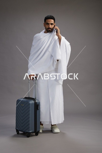 بورتريه لرجل عربي  سعودي خليجي بملابس الإحرام مع حقيبة سفر, استخدام الهاتف المحمول, الذهاب الى مكه للحج والعمره, كامل الجسم, خلفيه رماديه