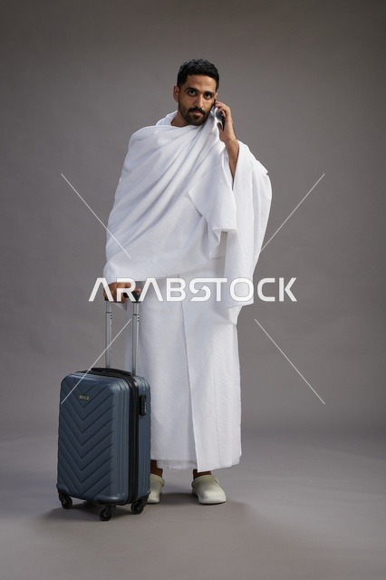 بورتريه لرجل عربي  سعودي خليجي بملابس الإحرام مع حقيبة سفر, استخدام الهاتف المحمول, الذهاب الى مكه للحج والعمره, كامل الجسم, خلفيه رماديه