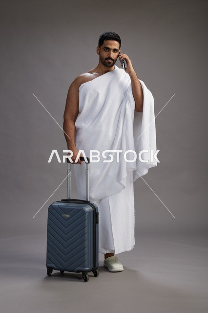 A portrait of an Arab Saudi Gulf man in Ihram clothing with a travel bag, using a mobile phone, going to Mecca for Hajj and Umrah, full body, gray background.