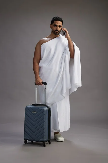 بورتريه لرجل عربي  سعودي خليجي بملابس الإحرام مع حقيبة سفر, استخدام الهاتف المحمول, الذهاب الى مكه للحج والعمره, كامل الجسم, خلفيه رماديه