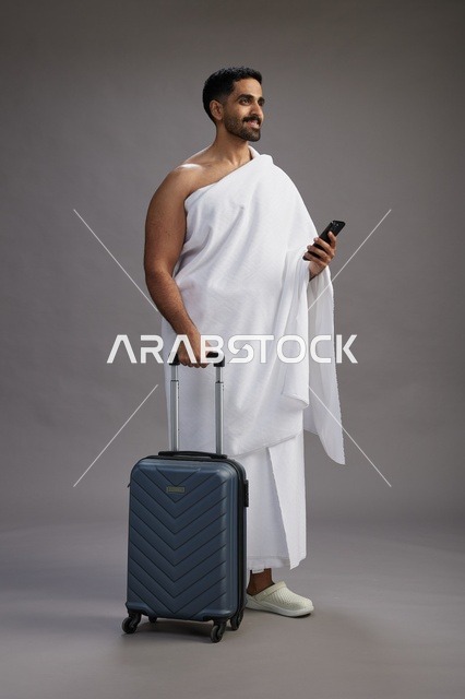 بورتريه لرجل عربي  سعودي خليجي بملابس الإحرام مع حقيبة سفر, استخدام الهاتف المحمول, الذهاب الى مكه للحج والعمره, كامل الجسم, خلفيه رماديه