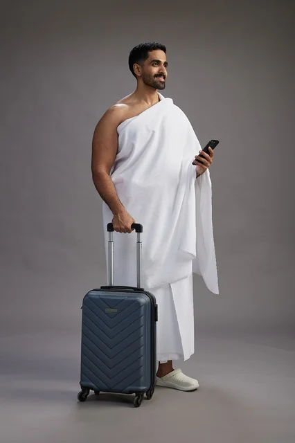 A portrait of an Arab Saudi Gulf man in Ihram clothing with a travel bag, using a mobile phone, going to Mecca for Hajj and Umrah, full body, gray background.