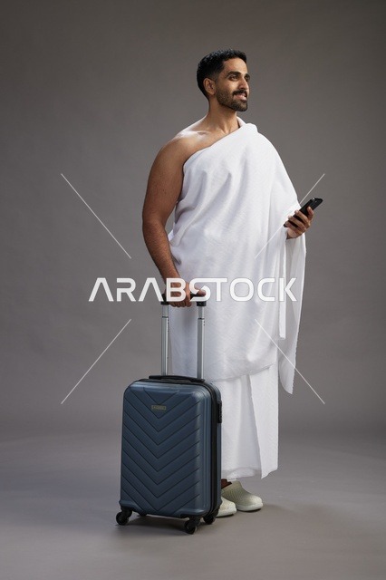 بورتريه لرجل عربي  سعودي خليجي بملابس الإحرام مع حقيبة سفر, استخدام الهاتف المحمول, الذهاب الى مكه للحج والعمره, كامل الجسم, خلفيه رماديه