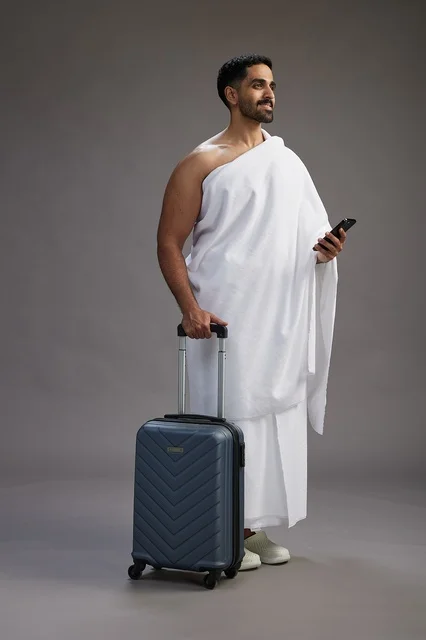 A portrait of an Arab Saudi Gulf man in Ihram clothing with a travel bag, using a mobile phone, going to Mecca for Hajj and Umrah, full body, gray background.
