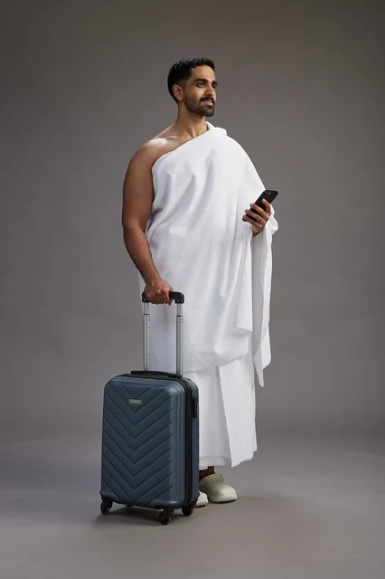 A portrait of an Arab Saudi Gulf man in Ihram clothing with a travel bag, using a mobile phone, going to Mecca for Hajj and Umrah, full body, gray background.
