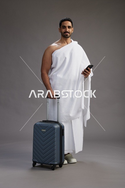 A portrait of an Arab Saudi Gulf man in Ihram clothing with a travel bag, using a mobile phone, going to Mecca for Hajj and Umrah, full body, gray background.