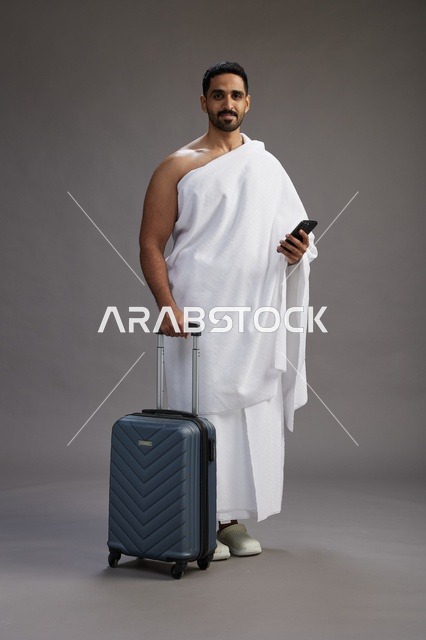 بورتريه لرجل عربي  سعودي خليجي بملابس الإحرام مع حقيبة سفر, استخدام الهاتف المحمول, الذهاب الى مكه للحج والعمره, كامل الجسم, خلفيه رماديه