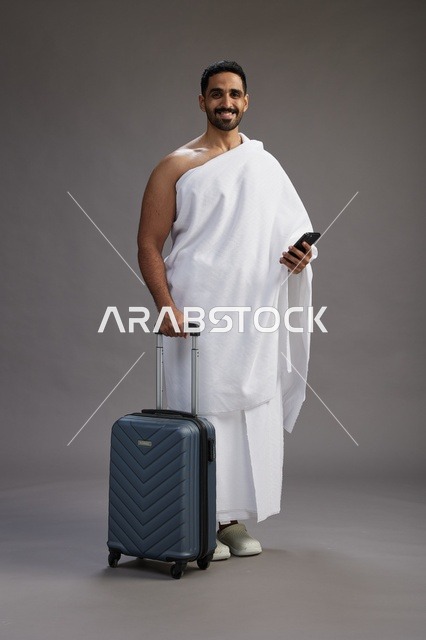 بورتريه لرجل عربي  سعودي خليجي بملابس الإحرام مع حقيبة سفر, استخدام الهاتف المحمول, الذهاب الى مكه للحج والعمره, كامل الجسم, خلفيه رماديه