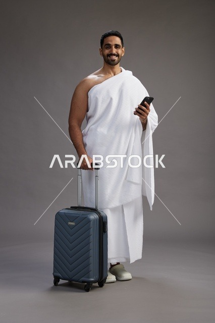بورتريه لرجل عربي  سعودي خليجي بملابس الإحرام مع حقيبة سفر, استخدام الهاتف المحمول, الذهاب الى مكه للحج والعمره, كامل الجسم, خلفيه رماديه