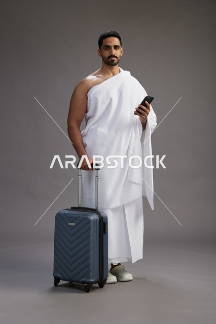 بورتريه لرجل عربي  سعودي خليجي بملابس الإحرام مع حقيبة سفر, استخدام الهاتف المحمول, الذهاب الى مكه للحج والعمره, كامل الجسم, خلفيه رماديه