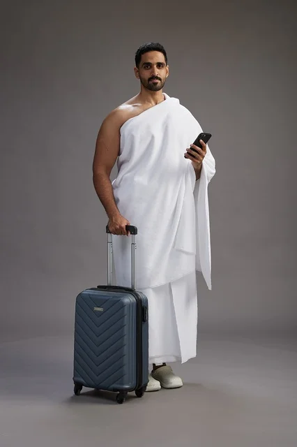 A portrait of an Arab Saudi Gulf man in Ihram clothing with a travel bag, using a mobile phone, going to Mecca for Hajj and Umrah, full body, gray background.