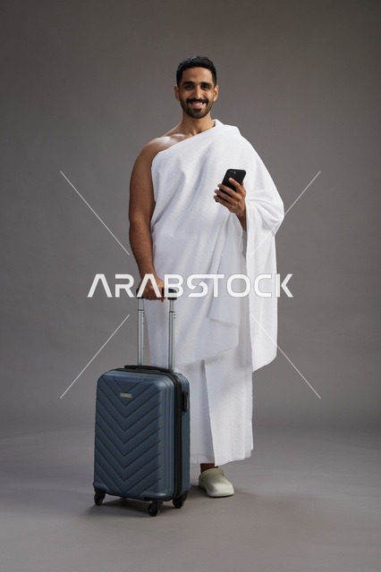 بورتريه لرجل عربي  سعودي خليجي بملابس الإحرام مع حقيبة سفر, استخدام الهاتف المحمول, الذهاب الى مكه للحج والعمره, كامل الجسم, خلفيه رماديه