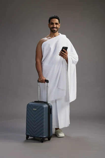 A portrait of an Arab Saudi Gulf man in Ihram clothing with a travel bag, using a mobile phone, going to Mecca for Hajj and Umrah, full body, gray background.