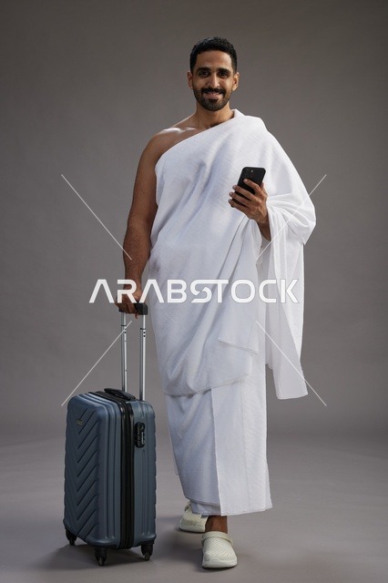 بورتريه لرجل عربي  سعودي خليجي بملابس الإحرام مع حقيبة سفر, استخدام الهاتف المحمول, الذهاب الى مكه للحج والعمره, كامل الجسم, خلفيه رماديه