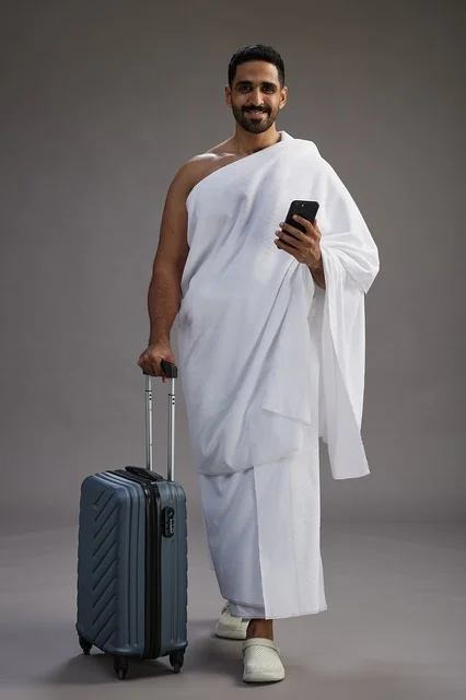 A portrait of an Arab Saudi Gulf man in Ihram clothing with a travel bag, using a mobile phone, going to Mecca for Hajj and Umrah, full body, gray background.