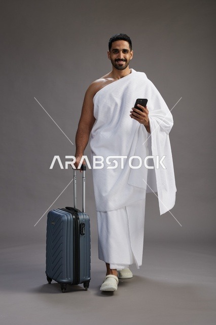 بورتريه لرجل عربي  سعودي خليجي بملابس الإحرام مع حقيبة سفر, استخدام الهاتف المحمول, الذهاب الى مكه للحج والعمره, كامل الجسم, خلفيه رماديه