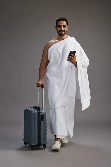 A portrait of an Arab Saudi Gulf man in Ihram clothing with a travel bag, using a mobile phone, going to Mecca for Hajj and Umrah, full body, gray background.