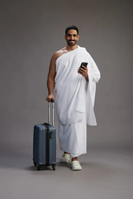 بورتريه لرجل عربي  سعودي خليجي بملابس الإحرام مع حقيبة سفر, استخدام الهاتف المحمول, الذهاب الى مكه للحج والعمره, كامل الجسم, خلفيه رماديه
