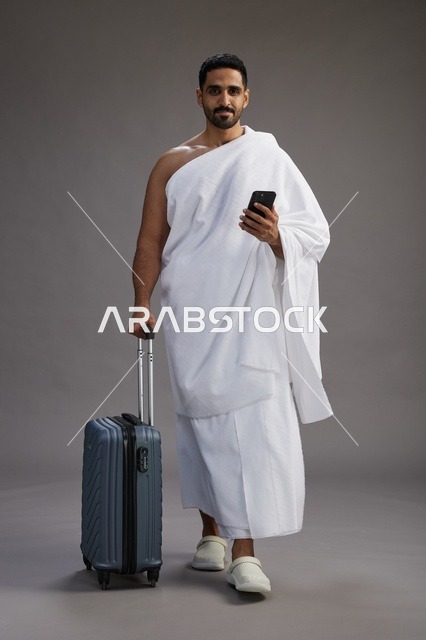 بورتريه لرجل عربي  سعودي خليجي بملابس الإحرام مع حقيبة سفر, استخدام الهاتف المحمول, الذهاب الى مكه للحج والعمره, كامل الجسم, خلفيه رماديه