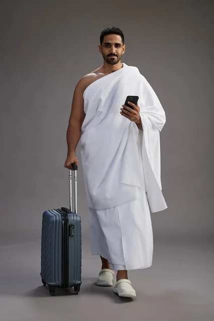 A portrait of an Arab Saudi Gulf man in Ihram clothing with a travel bag, using a mobile phone, going to Mecca for Hajj and Umrah, full body, gray background.