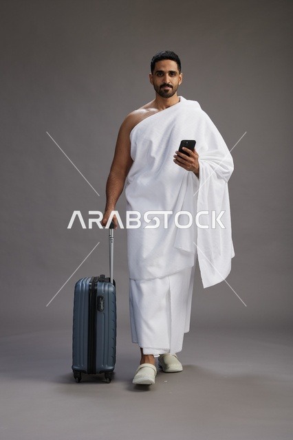 بورتريه لرجل عربي  سعودي خليجي بملابس الإحرام مع حقيبة سفر, استخدام الهاتف المحمول, الذهاب الى مكه للحج والعمره, كامل الجسم, خلفيه رماديه