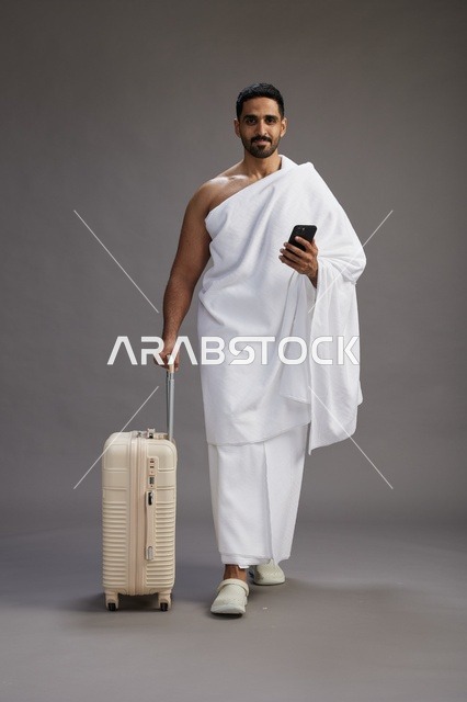 بورتريه لرجل عربي  سعودي خليجي بملابس الإحرام مع حقيبة سفر, استخدام الهاتف المحمول, الذهاب الى مكه للحج والعمره, كامل الجسم, خلفيه رماديه