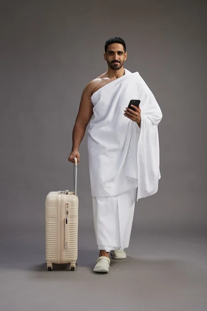 A portrait of an Arab Saudi Gulf man in Ihram clothing with a travel bag, using a mobile phone, going to Mecca for Hajj and Umrah, full body, gray background.