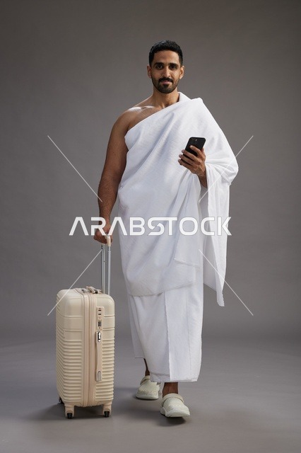 بورتريه لرجل عربي  سعودي خليجي بملابس الإحرام مع حقيبة سفر, استخدام الهاتف المحمول, الذهاب الى مكه للحج والعمره, كامل الجسم, خلفيه رماديه