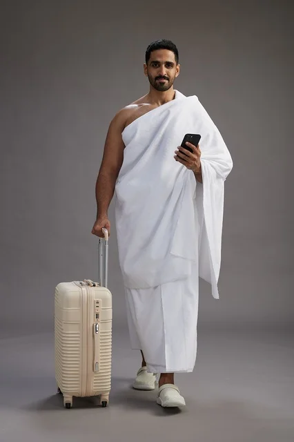 A portrait of an Arab Saudi Gulf man in Ihram clothing with a travel bag, using a mobile phone, going to Mecca for Hajj and Umrah, full body, gray background.