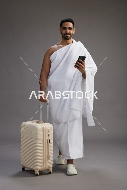 بورتريه لرجل عربي  سعودي خليجي بملابس الإحرام مع حقيبة سفر, استخدام الهاتف المحمول, الذهاب الى مكه للحج والعمره, كامل الجسم, خلفيه رماديه