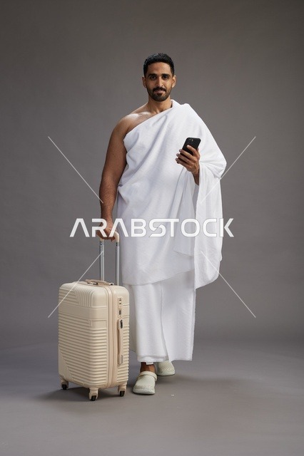 بورتريه لرجل عربي  سعودي خليجي بملابس الإحرام مع حقيبة سفر, استخدام الهاتف المحمول, الذهاب الى مكه للحج والعمره, كامل الجسم, خلفيه رماديه