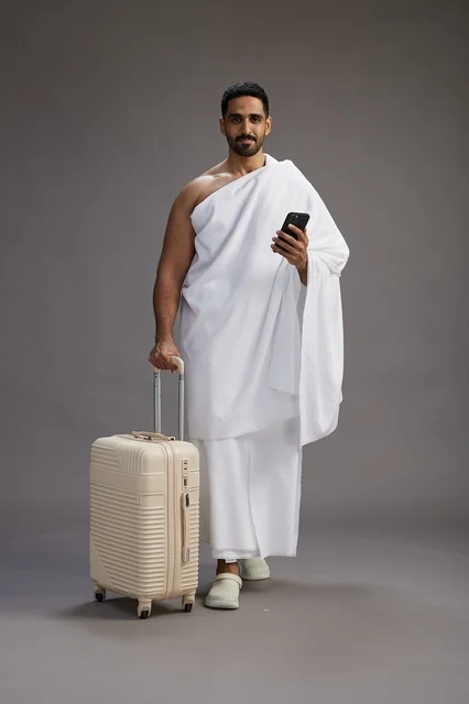 A portrait of an Arab Saudi Gulf man in Ihram clothing with a travel bag, using a mobile phone, going to Mecca for Hajj and Umrah, full body, gray background.