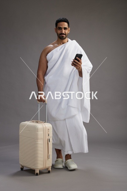 بورتريه لرجل عربي  سعودي خليجي بملابس الإحرام مع حقيبة سفر, استخدام الهاتف المحمول, الذهاب الى مكه للحج والعمره, كامل الجسم, خلفيه رماديه