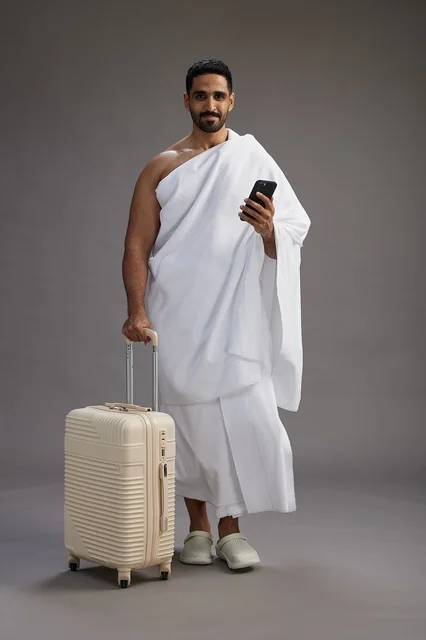 A portrait of an Arab Saudi Gulf man in Ihram clothing with a travel bag, using a mobile phone, going to Mecca for Hajj and Umrah, full body, gray background.