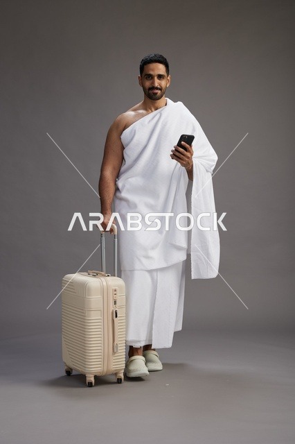 بورتريه لرجل عربي  سعودي خليجي بملابس الإحرام مع حقيبة سفر, استخدام الهاتف المحمول, الذهاب الى مكه للحج والعمره, كامل الجسم, خلفيه رماديه