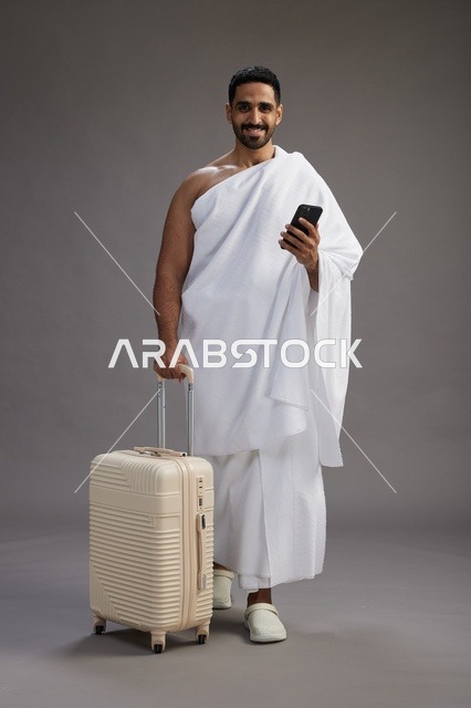 بورتريه لرجل عربي  سعودي خليجي بملابس الإحرام مع حقيبة سفر, استخدام الهاتف المحمول, الذهاب الى مكه للحج والعمره, كامل الجسم, خلفيه رماديه