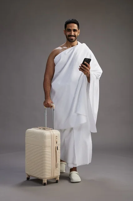 A portrait of an Arab Saudi Gulf man in Ihram clothing with a travel bag, using a mobile phone, going to Mecca for Hajj and Umrah, full body, gray background.