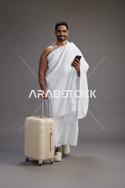 بورتريه لرجل عربي  سعودي خليجي بملابس الإحرام مع حقيبة سفر, استخدام الهاتف المحمول, الذهاب الى مكه للحج والعمره, كامل الجسم, خلفيه رماديه