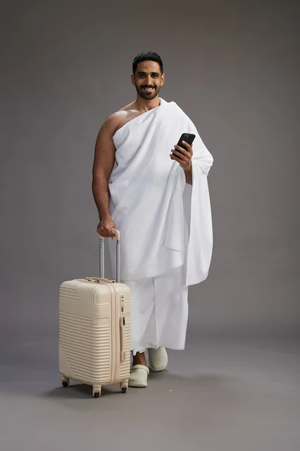 بورتريه لرجل عربي  سعودي خليجي بملابس الإحرام مع حقيبة سفر, استخدام الهاتف المحمول, الذهاب الى مكه للحج والعمره, كامل الجسم, خلفيه رماديه