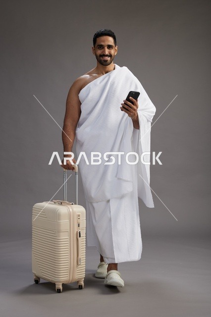 بورتريه لرجل عربي  سعودي خليجي بملابس الإحرام مع حقيبة سفر, استخدام الهاتف المحمول, الذهاب الى مكه للحج والعمره, كامل الجسم, خلفيه رماديه