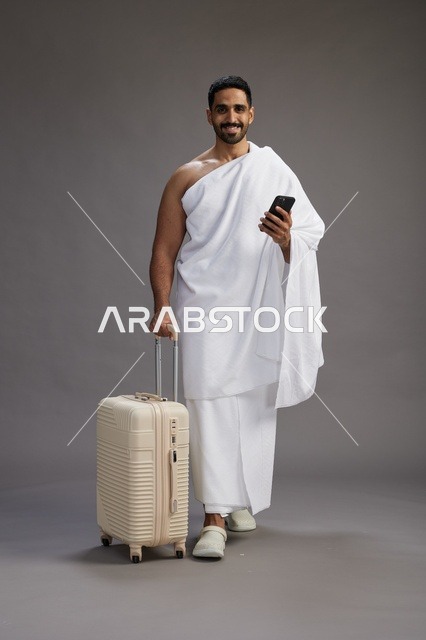 بورتريه لرجل عربي  سعودي خليجي بملابس الإحرام مع حقيبة سفر, استخدام الهاتف المحمول, الذهاب الى مكه للحج والعمره, كامل الجسم, خلفيه رماديه