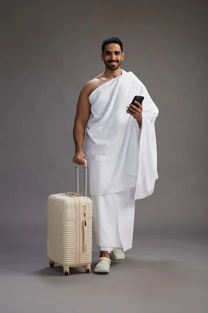 بورتريه لرجل عربي  سعودي خليجي بملابس الإحرام مع حقيبة سفر, استخدام الهاتف المحمول, الذهاب الى مكه للحج والعمره, كامل الجسم, خلفيه رماديه