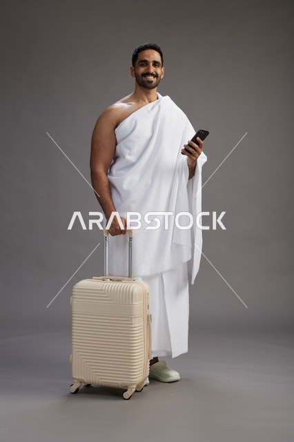 بورتريه لرجل عربي  سعودي خليجي بملابس الإحرام مع حقيبة سفر, استخدام الهاتف المحمول, الذهاب الى مكه للحج والعمره, كامل الجسم, خلفيه رماديه